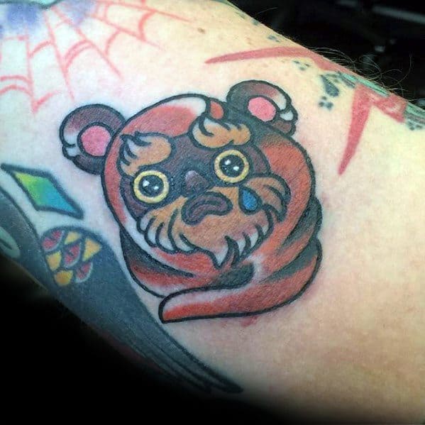 Cute and minimal Ewok tattoo with big yellow eyes and a single blue tear, wrapped in an orange hood on a colorful tattooed arm.