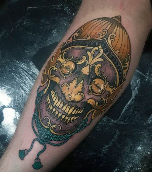 Tibetan skull tattoo on calf with golden helmet, purple shading, ornate details, and turquoise tassels at the base.