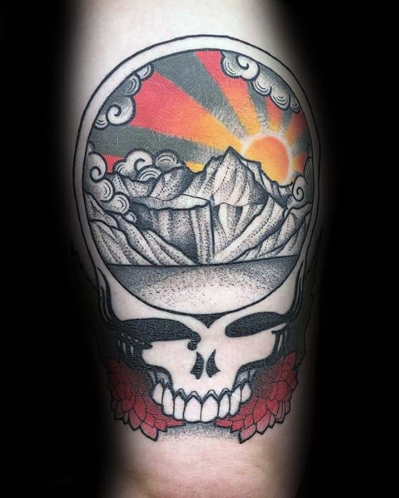 Grateful Dead Steal Your Face skull tattoo with dotwork mountains, sunrise rays, clouds, and red flowers at the base.