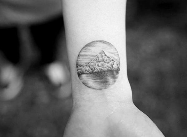 Small circular iceberg tattoo on the wrist with realistic shading, calm ocean reflection, and cloudy sky background.