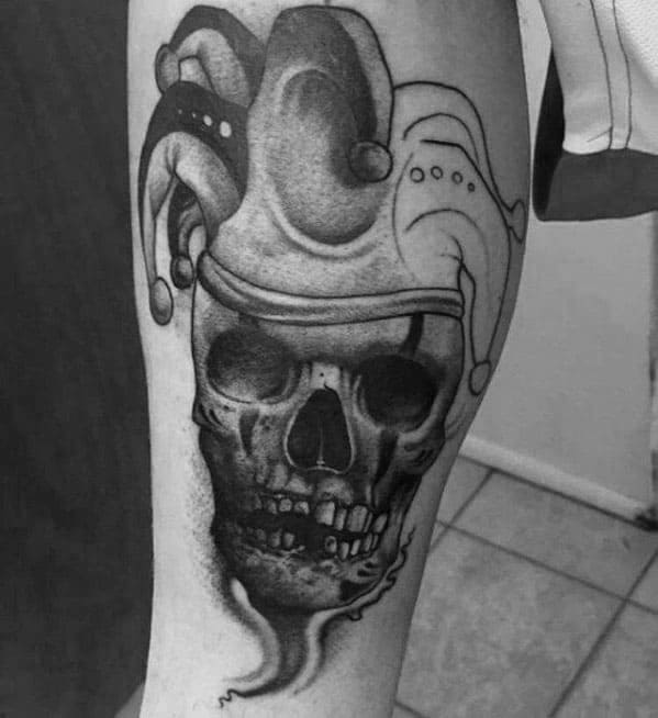 A black and gray tattoo of a skull wearing a jester hat, with intricate shading blending the skull and the playful jester details.