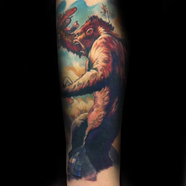 Vibrant full-color King Kong tattoo showing him atop a building, swatting planes mid-air with a dramatic sky in the background.