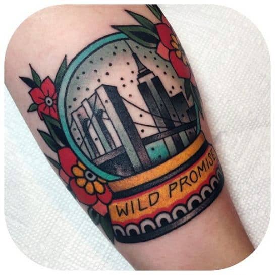 Colorful tattoo of New York City inside a snow globe with the Brooklyn Bridge, skyline, floral accents, and “Wild Promise” text.