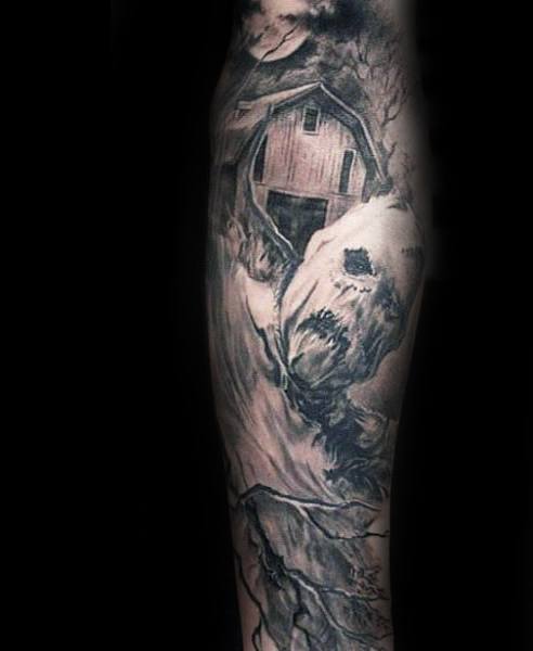 Dark scarecrow tattoo with a decaying face beneath a barn, surrounded by leafless trees and swirling shadows in a haunting blackwork style.