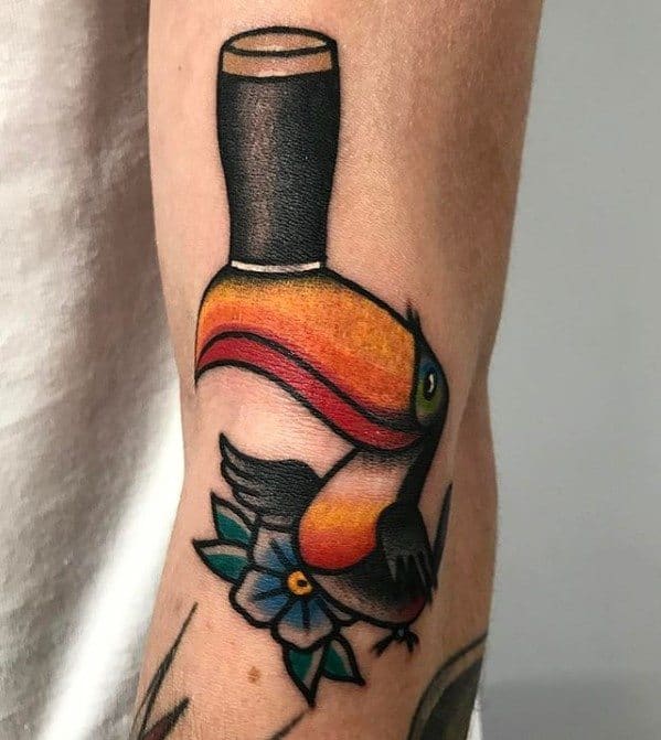 Traditional-style toucan tattoo with bold black outlines, a bright orange beak, and a blue flower accent on the forearm.