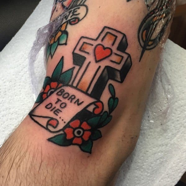 Traditional-style tattoo of a cross with a red heart, surrounded by flowers, leaves, and a scroll reading "BORN TO DIE..." on the forearm.