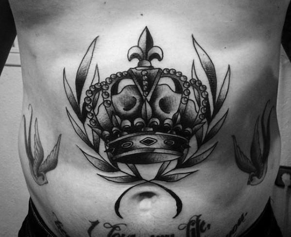 Black and grey crown tattoo on the stomach, detailed with laurel leaves and flanked by two birds in a traditional bold style.