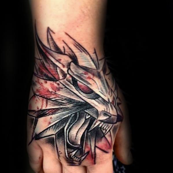 Bold Witcher wolf medallion tattoo on the hand with sharp lines, red splashes, and an aggressive, sketch-style design.