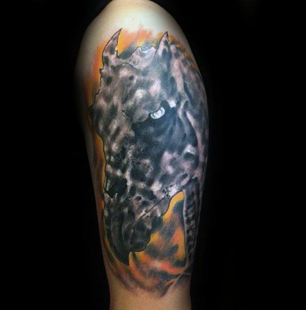 Realistic Thestral head tattoo with intense glowing eye and smoky textures, set against a fiery orange and black background on the upper arm.