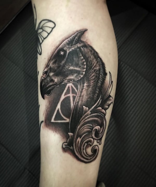Elegant Thestral head tattoo with flowing ornamental details and the Deathly Hallows symbol glowing in the background on the calf.
