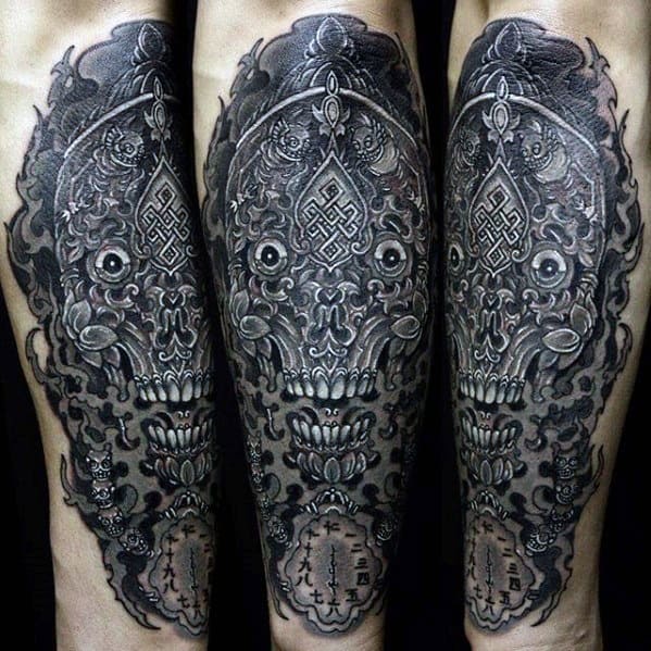 Dark Tibetan skull calf tattoo in black and grey, featuring intricate patterns, sacred symbols, and textured shading for a bold look.