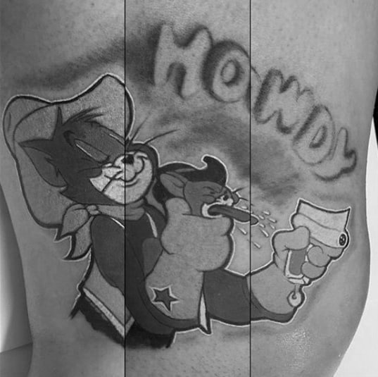 Black and grey tattoo of cowboy Tom punching Jerry while saying “Howdy,” done in a bold Western cartoon style.