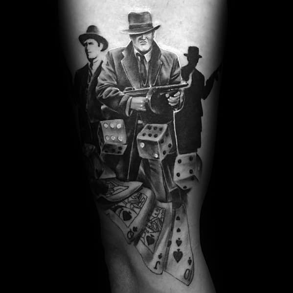 Black and grey gangster tattoo featuring a mobster holding a Tommy gun, surrounded by playing cards and dice, symbolizing crime and gambling.