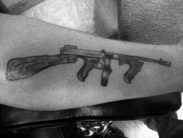 Realistic black and grey Tommy gun tattoo on the forearm, featuring fine details and shading for a classic firearm design with a vintage feel.