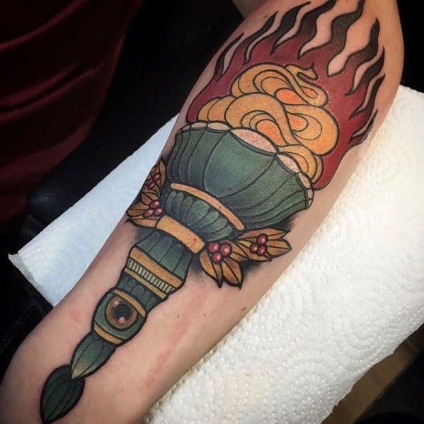 Traditional torch tattoo with bold black flames, golden smoke swirls, olive green handle, and red floral accents on the sides.