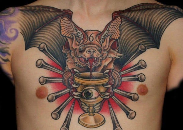 Large traditional bat tattoo on the chest with outstretched wings, holding a chalice with an eye and surrounded by radiating bones.