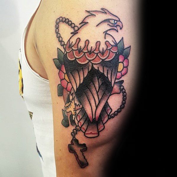 Traditional eagle tattoo on the upper arm with flowers and a rosary wrapped around it, ending in a cross hanging below the wing.