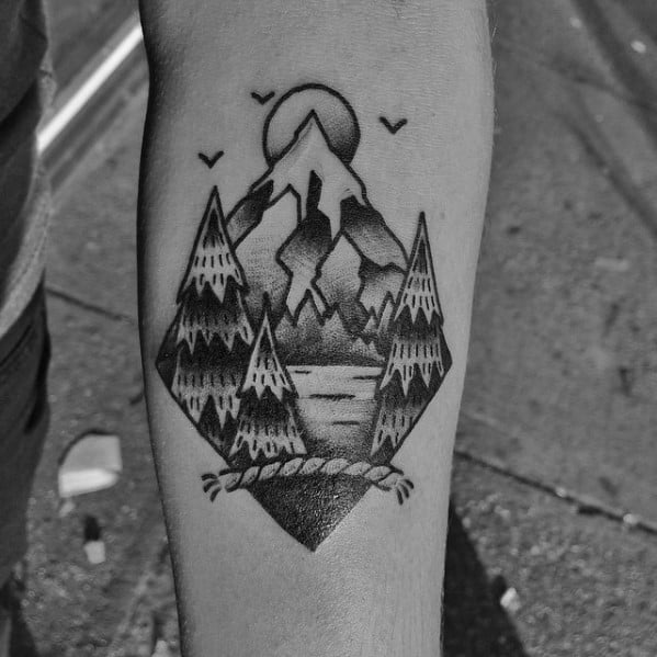 Black and grey tattoo of pine trees, a mountain peak, and a lake, framed in a diamond with a rope knot at the bottom.