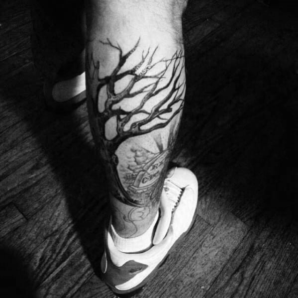 Black ink tree tattoo on the leg, featuring twisted branches extending around the calf, blending with intricate clock and eye elements.