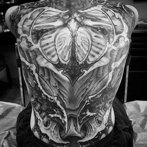 Dark biomechanical full-back tattoo in black and grey, featuring rib-like structures, layered textures, and a symmetrical, alien-inspired design.