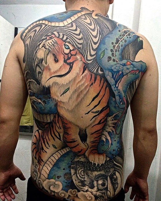 Dynamic full-back tattoo of a fierce tiger standing over a stylized face, with swirling blue patterns and a serpent-like creature behind it.