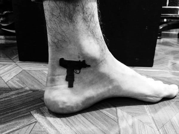 Small black Uzi tattoo on the ankle, featuring a minimalist silhouette design with bold ink contrast.