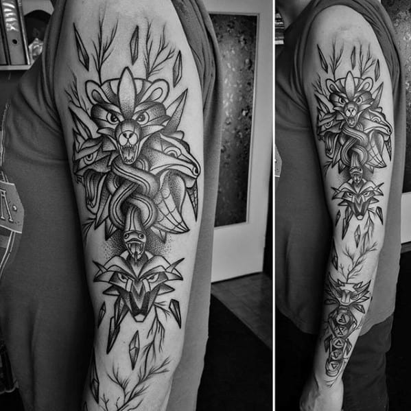 Black and grey full sleeve Witcher tattoo featuring a stylized wolf medallion, snakes, and swords in a bold geometric design.