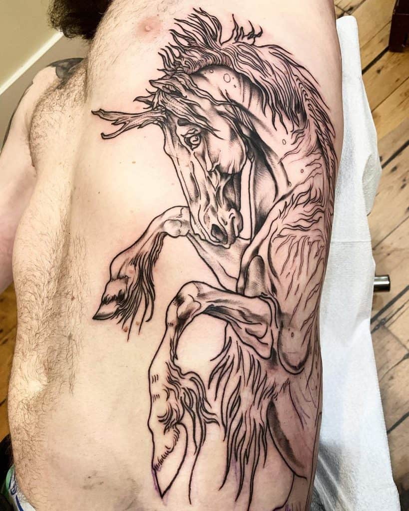 Large black ink tattoo of a fierce rearing unicorn with bold linework and intricate details, covering the ribcage and side torso.