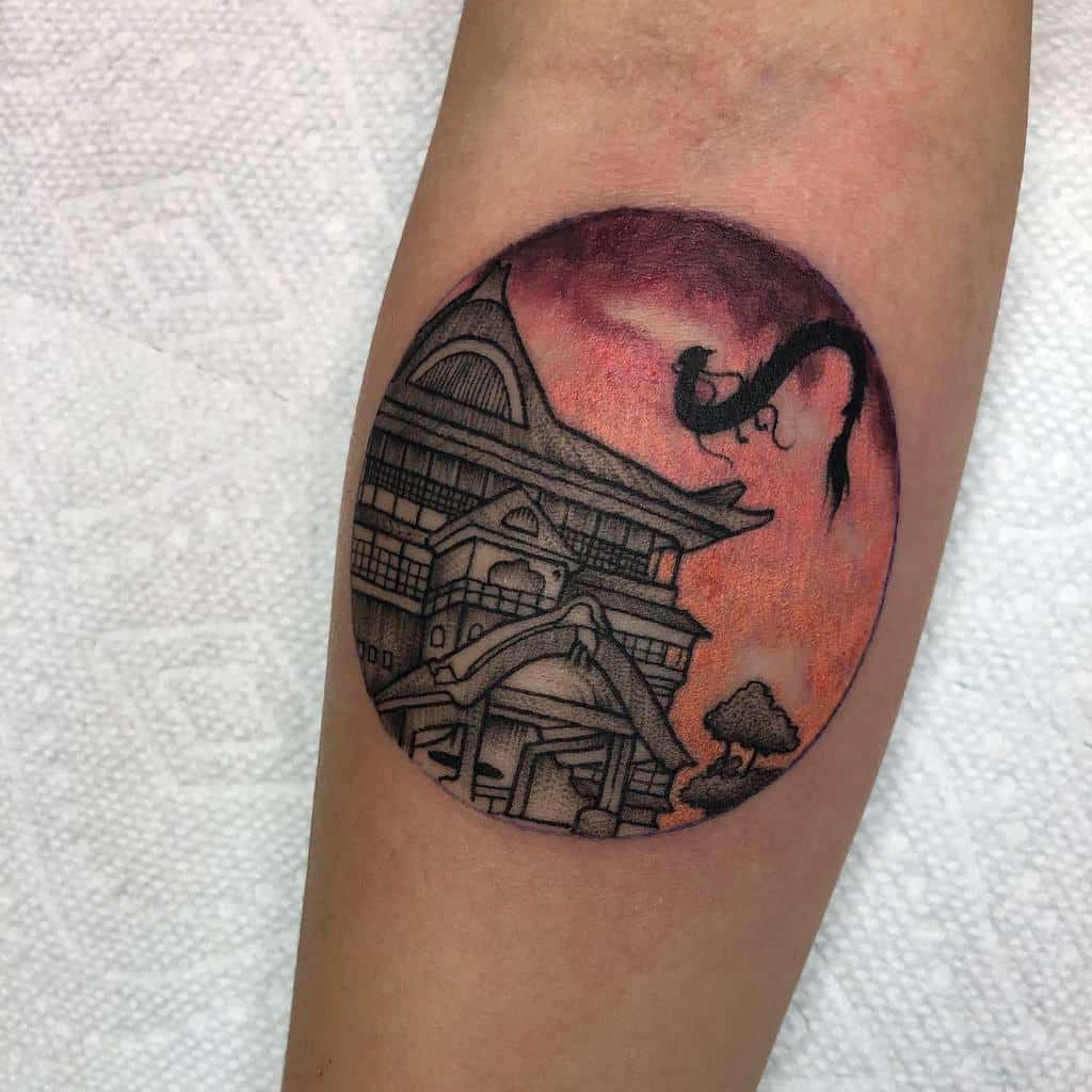 Stunning Spirited Away tattoo of the bathhouse with Haku in dragon form soaring against a warm sunset sky, framed within a circular design