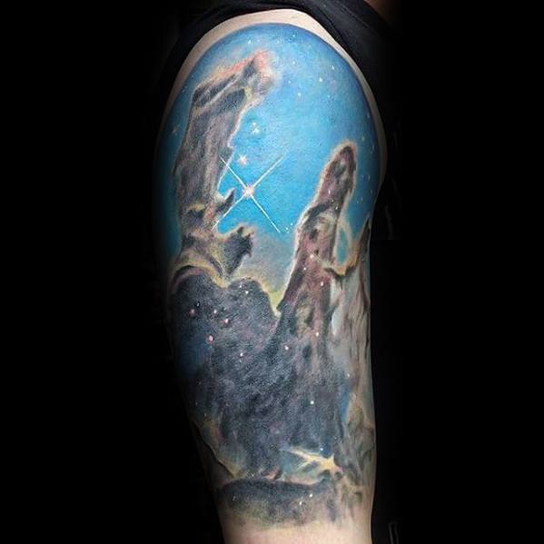 Nebula tattoo of the Pillars of Creation on an upper arm, featuring bright stars and intricate nebula details