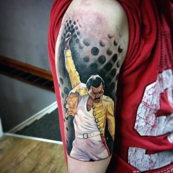 Vibrant Freddie Mercury tattoo in yellow jacket with raised fist, set against a bold black dotwork background on the upper arm.