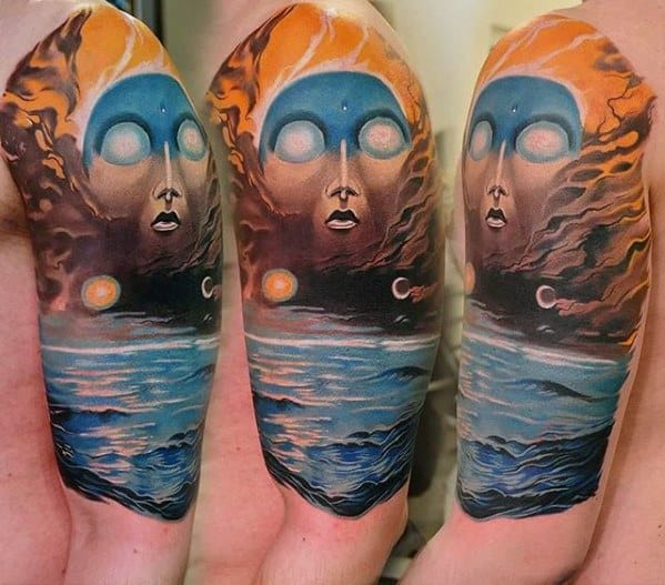 Surreal tattoo of a glowing-eyed face emerging from the sky above a cosmic ocean, blending celestial elements with dreamlike waves.