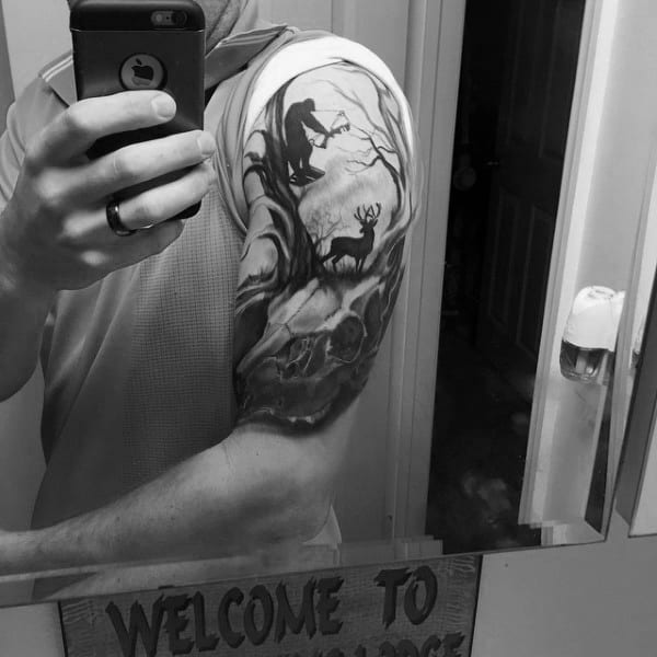 Black and grey upper arm tattoo of a bowhunter aiming at a deer in a forest scene, with detailed trees and a deer skull below.