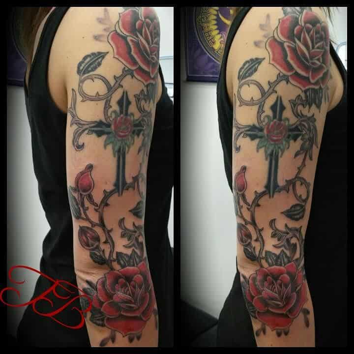 A half-sleeve tattoo featuring bold red roses intertwined with dark vines and a cross, blending vibrant colors with intricate detailing for a dramatic design