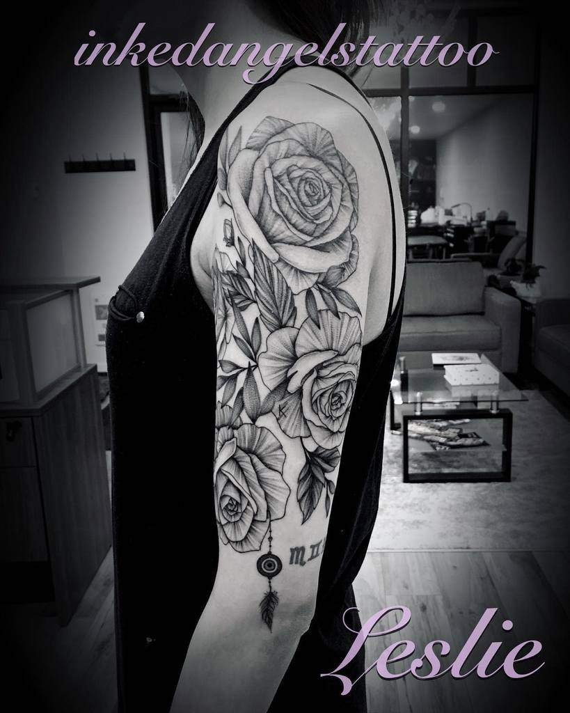 A beautiful half-sleeve tattoo featuring detailed black and gray roses, with intricate shading and soft highlights, accompanied by a feather and an evil eye charm hanging from the design