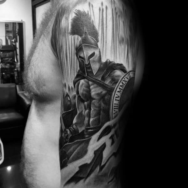 Black and grey upper arm tattoo of a muscular Spartan warrior with a plumed helmet, holding a shield engraved with Roman numerals.