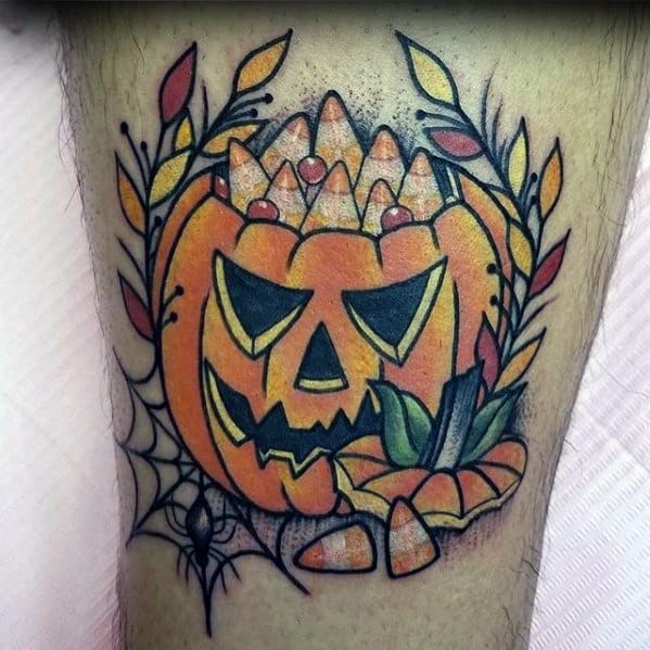 Bold Halloween-themed tattoo of a sinister jack-o’-lantern filled with candy corn, surrounded by autumn leaves, a spiderweb, and a spider.