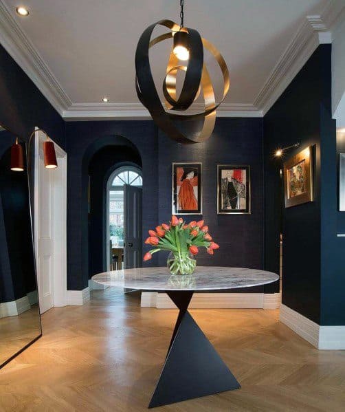 A stylish hallway with navy walls, elegant crown molding, a geometric chandelier, and a modern table with fresh flowers