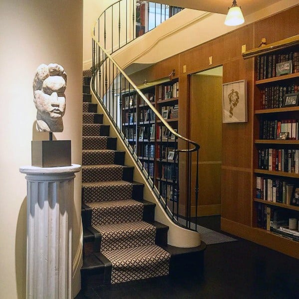 Elegant hallway with built-in bookcases, spiral staircase, patterned carpet, and artistic bust on a pedestal