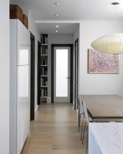 Minimalist hallway with a narrow built-in bookcase, light wood flooring, modern dining area, and abstract wall art