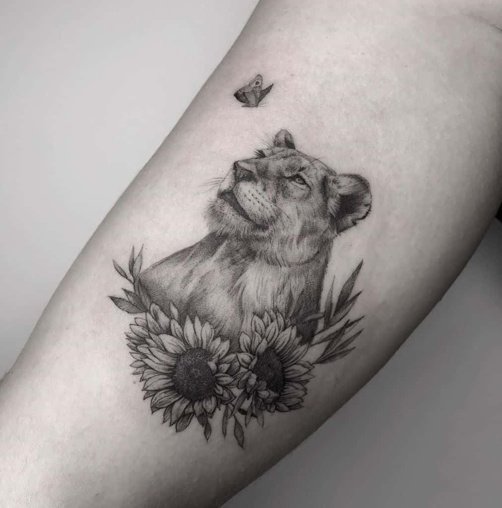 Black and grey lioness tattoo on the forearm, featuring realistic shading, sunflowers, and a butterfly for a serene and powerful design.