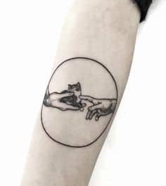 A black ink tattoo parodying 'The Creation of Adam,' replacing the central touch with a tiny cat reaching out, enclosed in a circular frame.