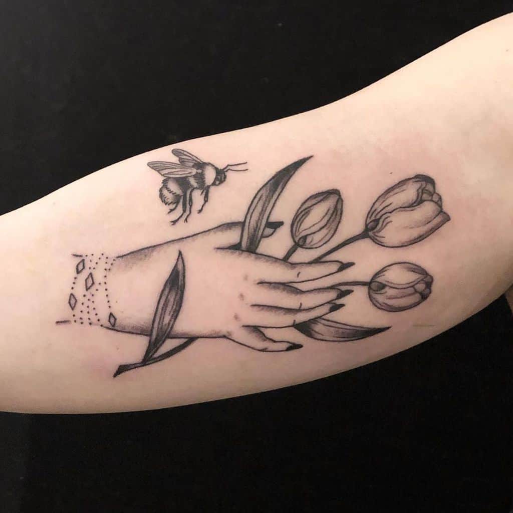 A black and grey tattoo featuring a hand reaching for three tulips, with a bee hovering above and delicate ornamental details on the wrist.