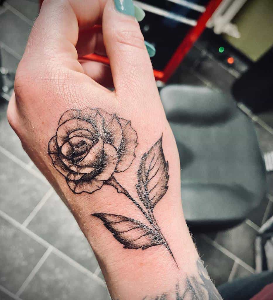 A detailed black and white tattoo of a rose on the hand, with shading and fine linework on the petals and leaves