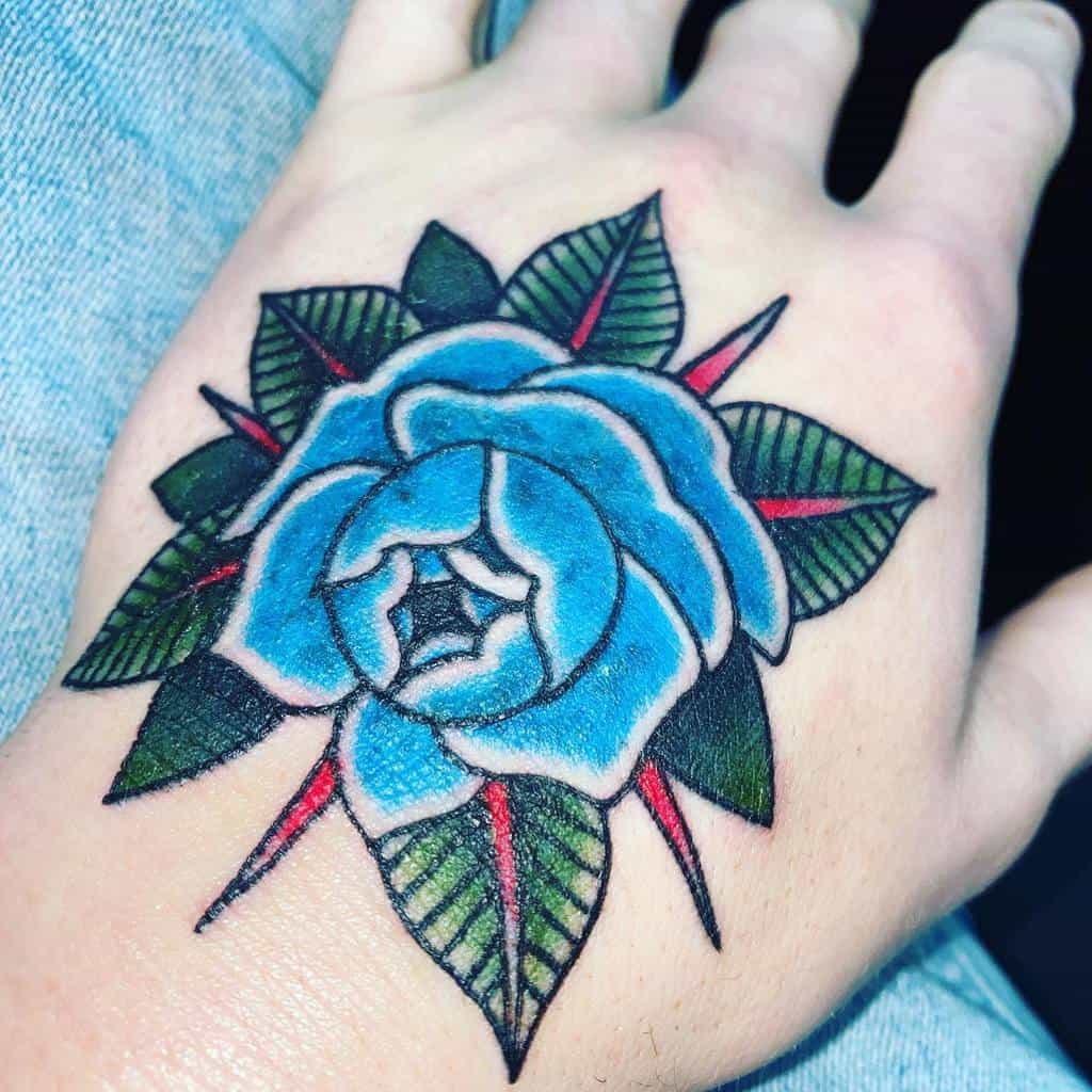 A stunning blue rose tattoo blooms gracefully on a person's hand, its vibrant green leaves and subtle red accents adding depth and allure to the design