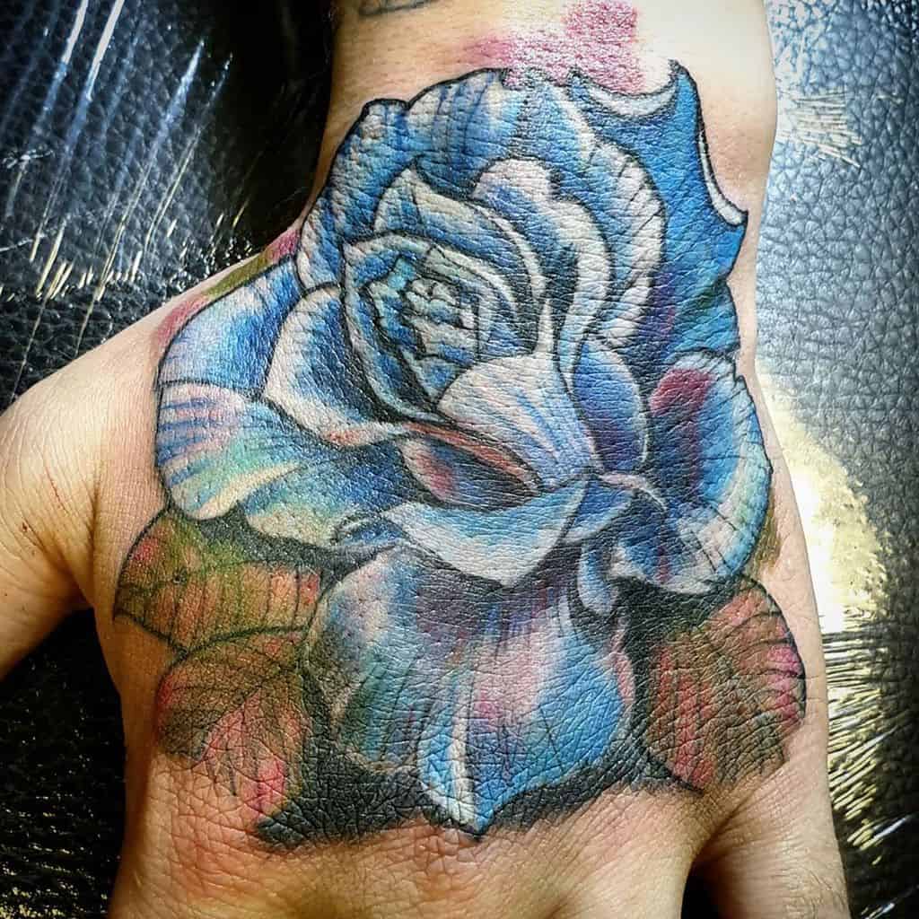 A vibrant, multicolored blue rose tattoo on the hand, featuring detailed petals and shading with hints of pink, purple, and green for depth