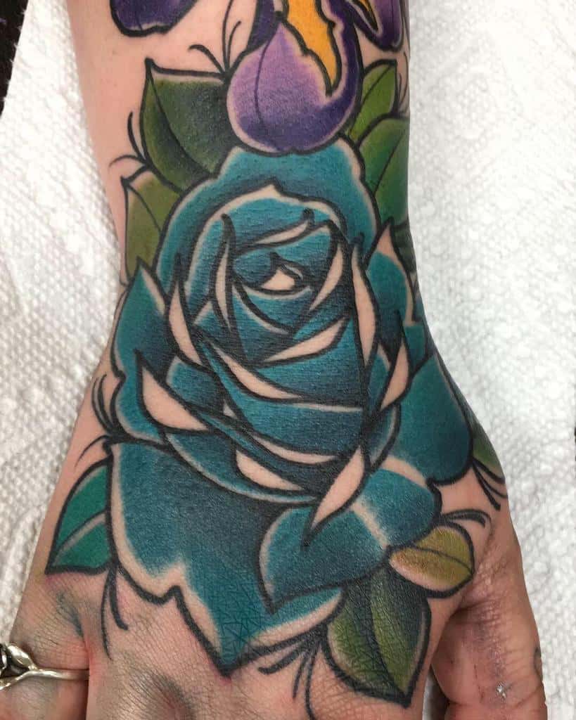 A vibrant hand tattoo of a blue rose intertwined with a delicate purple flower, all set against lush green leaves