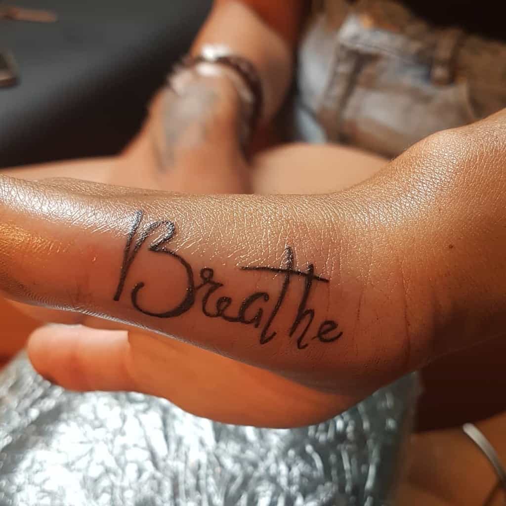 A tattooed hand showcases the word "Breathe," gracefully scripted along the inside edge of the thumb, a subtle yet powerful reminder to stay present