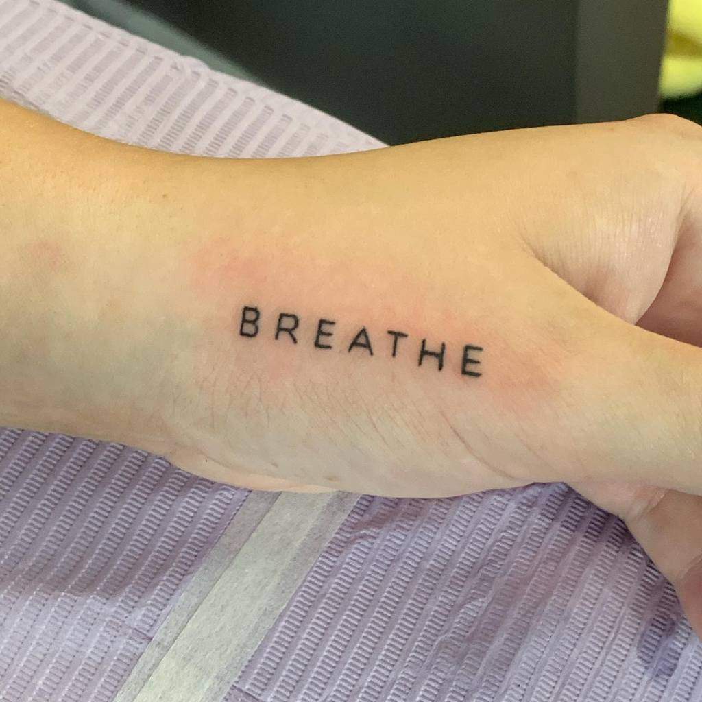 A wrist tattoo elegantly displays the word "BREATHE" in bold black capital letters, serving as a constant reminder to stay calm and centered