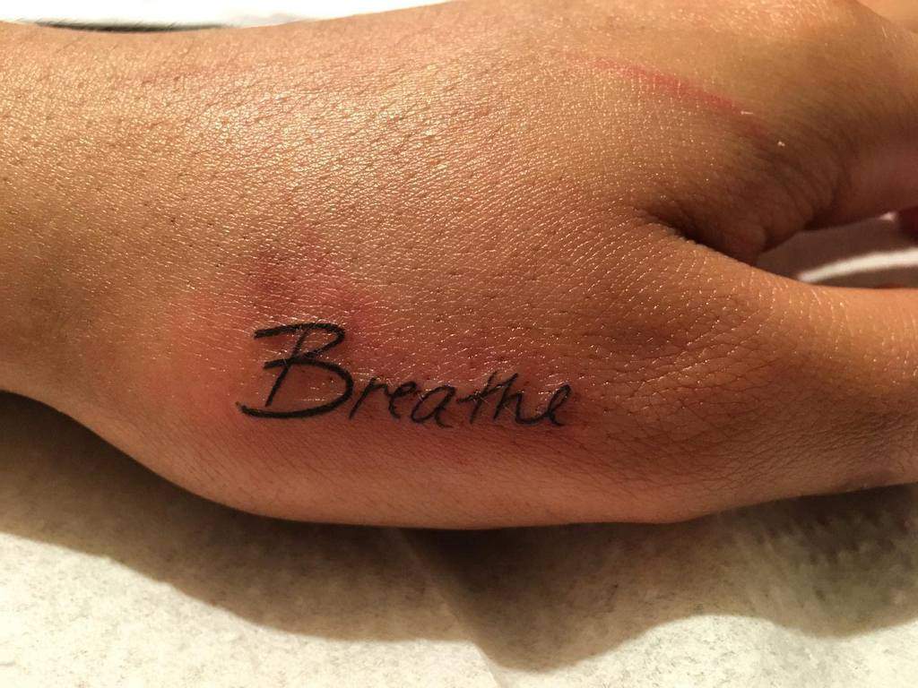 The back of the hand bears a delicate tattoo of "Breathe," serving as a subtle reminder to embrace tranquility in every moment
